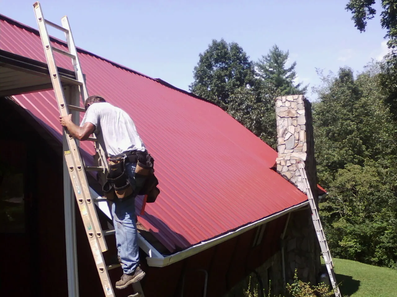 Roofer on a ladder beside a red metal roof and stone chimney — Soffit & Fascia Repair by Roofing Brothers in Manly